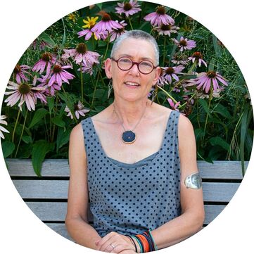 Photo of Lorraine Johnson smiling and sitting on a bench with blooming purple coneflowers behind. She is a white woman with very short silver hair, dangling earrings, round red glasses, a large blue floral pendant necklace, silver upper arm band and loose blue top.