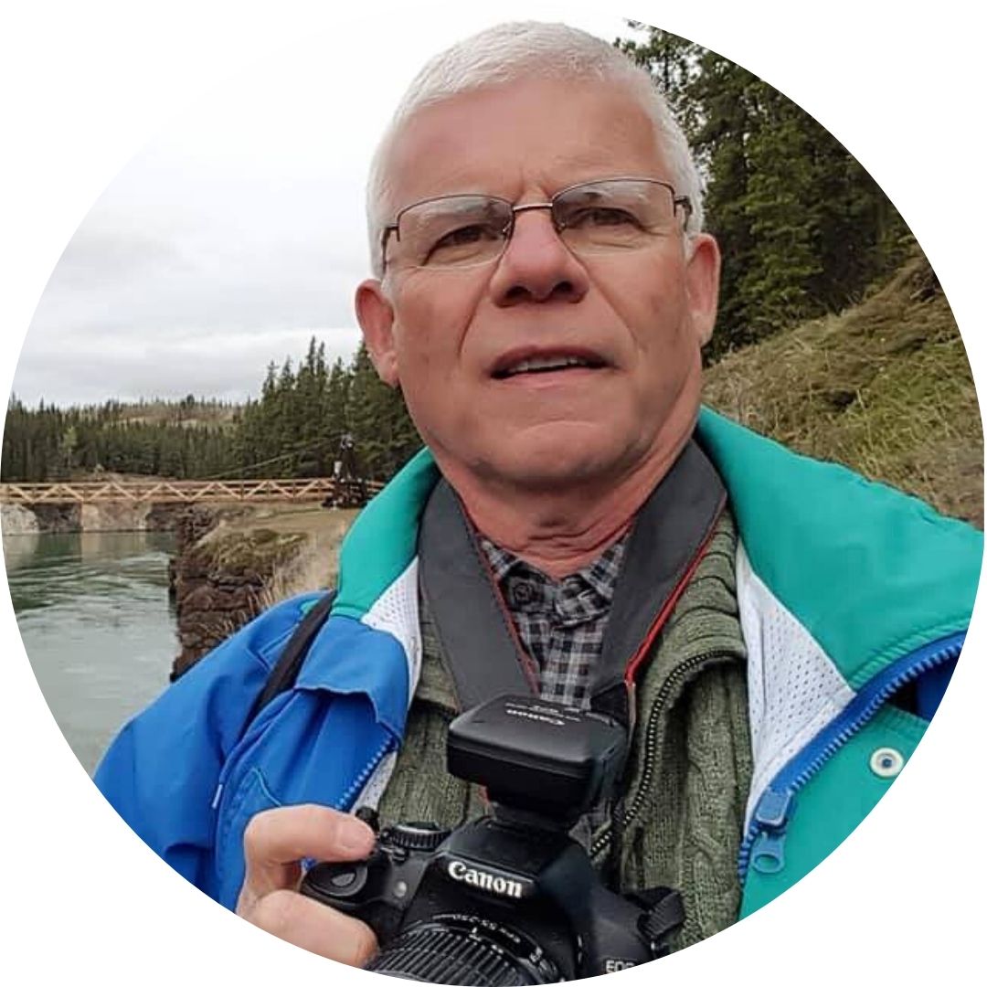 Photo of Rick Gray, white man with white hair and eyebrows, wearing glasses, holding a Canon camera. Background is a riverbank with tall trees.